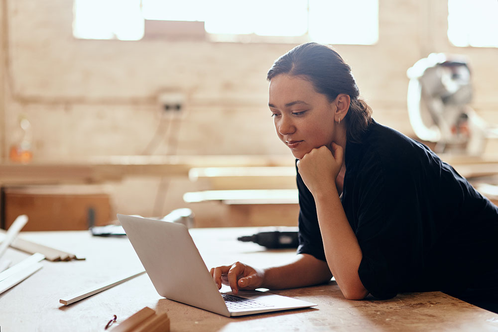 A young woman works on her laptop inside a carpenter's shop